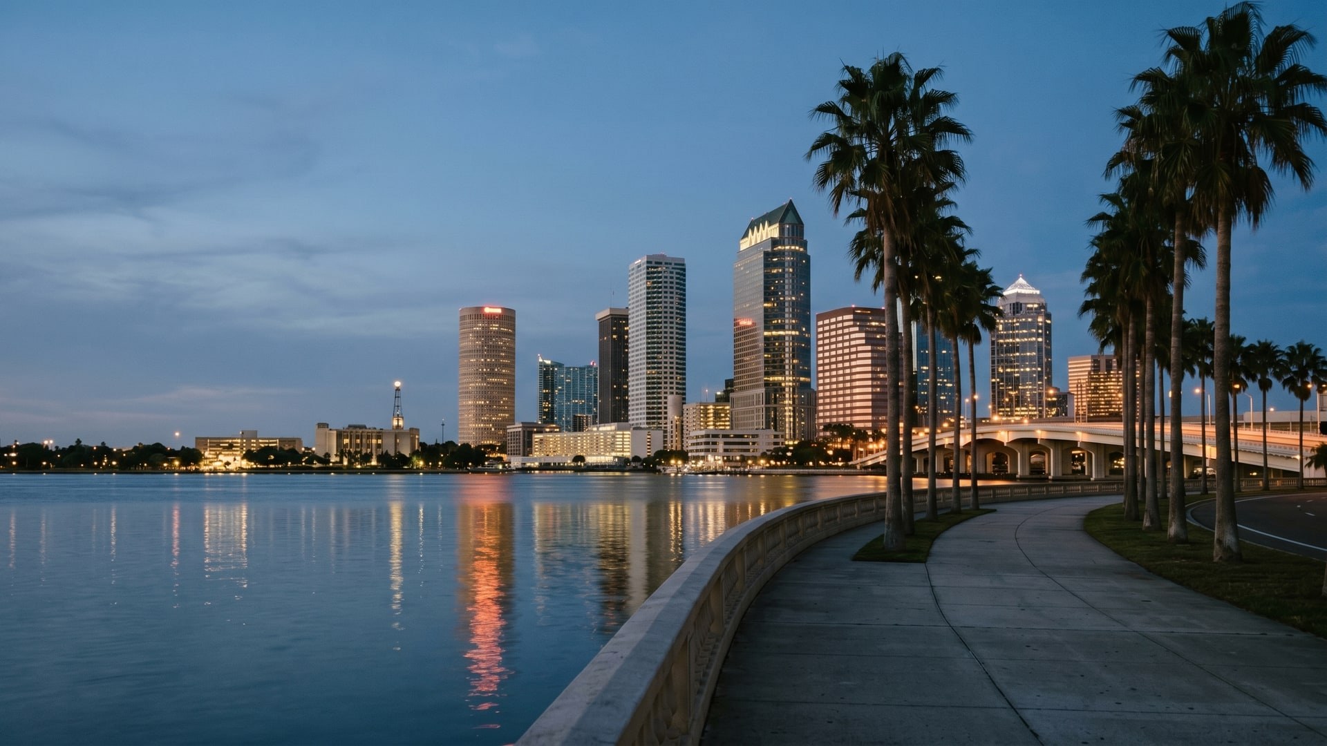 Downtown Tampa skyline reflecting in Hillsborough Bay at twilight, with the curving palm-lined Bayshore Boulevard in the foreground